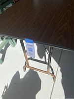 Close-up photo of the table corner showing woodgrain laminate top and metal folding leg structure.