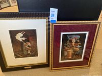 Two framed surrealist prints side by side on carpeted floor. Left with brown frame and cream mat, right with gold frame and burgundy mat.