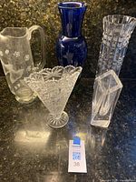 Whole five-piece set showing all items standing together on countertop: one tall blue glass etched vase, one clear pitcher with floral and bird motifs, one cut crystal vase, one hexagonal clear vase, one fan-shaped clear etched vase.