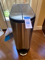 Front view of stainless steel rectangular trash can with foot pedal, positioned by door with broom and dust pan behind it.