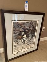 Full view of framed art print of merganser ducks leaning against wall, showing dark wooden frame and matting around print.