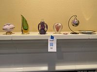 Full view of six collectible decorative items displayed on a white mantel including floral trinket box, glass art pieces, heart, hanging ornament, candy-shaped glass, and decorative plate.
