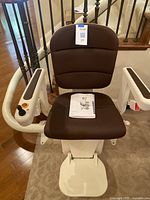 Front view of stairlift chair with brown seat, armrests with control joystick, and footrest; manual placed on seat.