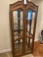 Front view of the wooden display cabinet with glass doors, showcasing the ornate carved detailing on the upper front.