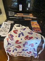 Wide shot showing apron, towels, grill grate, grill basket, utensils, and cookbook arranged on black countertop.