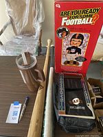 Overall view of items on table including bobblehead in box, football-related cup, wooden bats, and boxed gloves and visor.