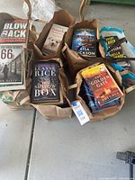 Multiple brown paper bags each containing paperback books, laid on the ground. Titles like 'Blowback', 'The Shadow Box', and 'The Golden Gate' are visible along with others.