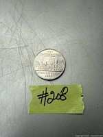 Photo of a silver-colored one dollar Canadian coin commemorating the Confederation Constitution from 1867 to 1982, placed on a scratched surface with a small piece of green tape labeled '#208'.