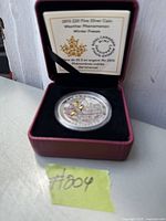 Photo of 2015 $20.00 fine silver coin inside its maroon display case with black velvet interior. Coin has colored elements representing winter freeze weather phenomenon. Informational insert visible in lid.