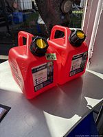 Two new red Scepter gas cans, each 5L capacity, with black caps and yellow spouts, displayed outdoors on concrete surface.