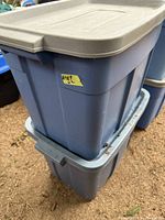Two stacked blue Roughneck Rubbermaid storage totes with tan lids on outdoor ground surface with dirt.