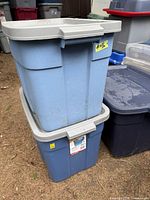 Two stacked Roughneck Rubbermaid totes outdoors, blue plastic with beige lids showing normal wear and exterior dirt marks.