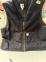 Front view of black Carhartt vest showing zip closure, front pockets with logo and Sherpa lining inside