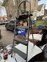 Photo of a folding step ladder with four steps and curved handle, resting outdoors on a table surface among other miscellaneous items.