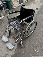 Full view of the folding wheelchair showing chrome metal frame, black vinyl seat and armrests, large rear wheels and footrests.
