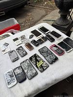 Wide view showing all cell phones and accessories arranged on a cloth surface outdoors. Various smartphones and older phone models, some with visible screen damage.