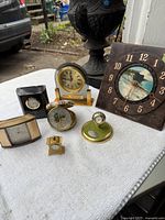 Overview of the lot showing seven clocks of varied sizes and styles on a white surface outdoors: includes world map clock, rectangular clock, scenic design clock, and small decorative clocks.