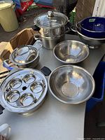 Overview of the lot showing stainless steel pots, bowls, a multi-tier steamer, and colanders on a table.