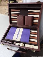 Photo of open portable backgammon case showing game board with brown and beige triangular points, two brown dice cups, dice, doubling cube, playing pieces, and instruction booklet.