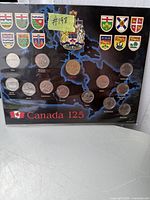 Display board showing a map of Canada with twelve coins placed according to their respective provinces and territories. Each coin is labeled with the name of the province or territory it represents, and above each coin is an emblem or shield representing that province or territory. The bottom of the board reads 'Canada 125'.
