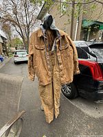 Full view of the brown Dickies jacket and overalls displayed together on a hanger outside near vehicles and chair