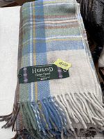 Folded wool blanket with tag showing the Highland Tartan Tweeds brand and fringe tassels at the edge.