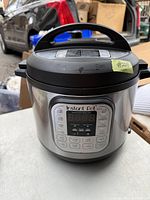 Front and top view of Instant Pot stainless steel pressure cooker with digital interface and black handle in an outdoor setting.