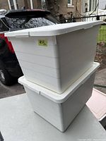 Two white plastic storage totes with fitted lids stacked, shown outside next to a car