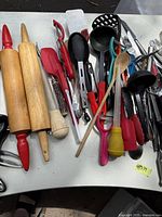 Assorted kitchen utensils including two wooden rolling pins with red handles, spatulas, tongs, peelers, wooden spoon, and ladles laid out on a table.