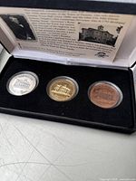 Photo of the set of three commemorative coins in presentation box, showing silver, gold, and bronze-tone coins with engraved buildings representing Dundas, Ontario. Information card visible inside the box lid.