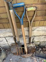 Three garden tools leaning against a brick wall showing a blue handled shovel, pitchfork, and other shovel handles with visible rust and wear