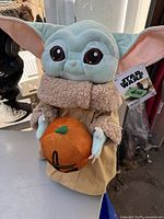 Plush figure of The Child (Grogu) from Star Wars dressed in a Halloween-themed outfit holding a pumpkin, showing the front and main features of the item.