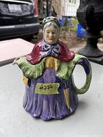 Photo showing a vintage ceramic teapot designed as an old woman wearing traditional clothing, including a bonnet and dress, predominantly purple and red colors.