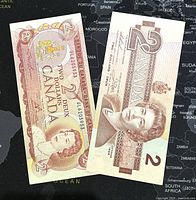 Two Canadian 2 dollar paper banknotes, one from 1974 and one from 1986, laid flat on a world map background showing front side details of both notes.