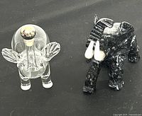 Two elephant figurines, one clear glass with rounded finial and another dark marbled stone with white tusks, displayed side-by-side on a black cloth.