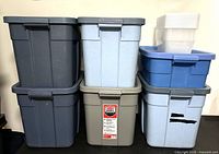 Six large Rubbermaid storage containers of various colors stacked against a wall.