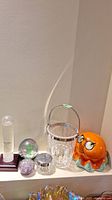 3 glass paperweights, crystal ice bucket, orange octopus coin bank, storm glass, and condiment jar on white shelf