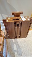 Top-front view of folded wooden artist's easel box showing leather handle and brass hardware latches