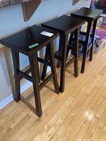 Front and side view of three black wooden bar stools lined up on wooden floor, showing seat tops and legs.