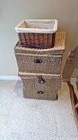 Stack of two rectangular woven seagrass storage boxes with leather straps, with a small dark brown wicker basket with fabric lining on top, against beige wall and carpeted floor.