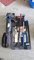 Overview of the lot on garage floor showing battery charger, creeper, various snow scrapers and brushes grouped on a black creeper.