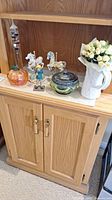 Wooden cabinet shelf with all items displayed: Galileo thermometer, two carousel horse figurines, art glass pumpkin, potpourri bowl with lid, ceramic pitcher vase with artificial roses.