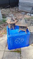 Two rusted cast iron pagoda-style candle holders placed on a blue recycling bin outdoors. One has a star-shaped roof, the other a pitched roof with a hole.