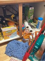 Under sink view showing multiple cleaning products, Swiffer Dusters box, roll paper towels, mop head, various spray bottles, and a green Swiffer vacuum.