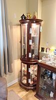Front view of the tall, curved glass curio cabinet with wooden frame, showing three glass shelves inside with some decorative items (contents not included in sale). Key lock is visible on the front door.
