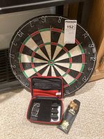 Full view of dart board leaning against furniture, black case open displaying darts, and separate black pouch containing brass-colored darts on carpet floor.