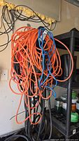 Tangled bundle of orange and black extension cords hanging on a garage wall near shelving.