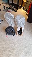 Four table fans on carpeted floor, showing variety in size and color including black and white fans. One fan is still boxed.