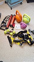 Fitness gear displayed including kettlebells, dumbbells, hand pump, resistance bands, and TRX Suspension Trainer straps on carpeted floor