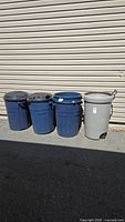 Five garbage cans lined against siding: four large blue cans with black lids and one smaller gray bin with wheels.